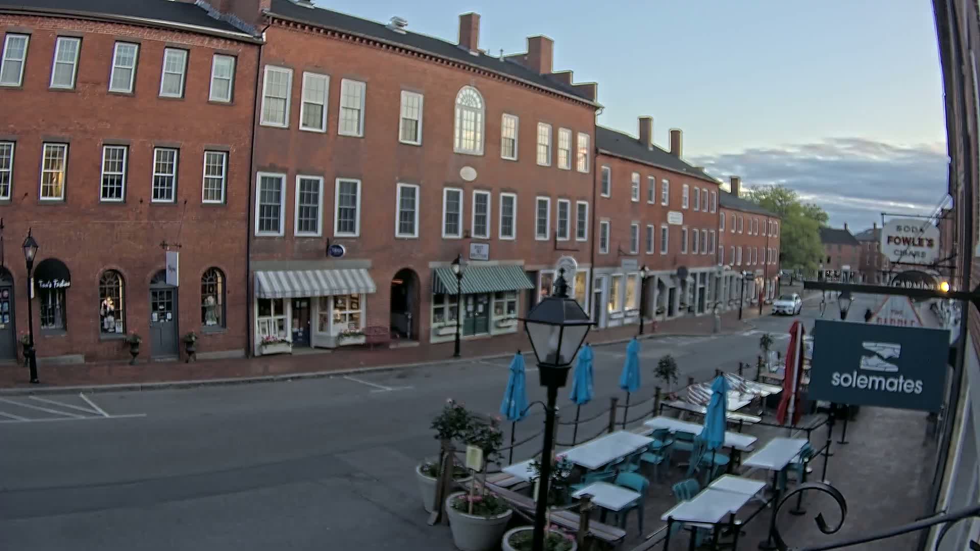 A brick street lined with shops and restaurants features outdoor seating with blue umbrellas under a partly cloudy evening sky.