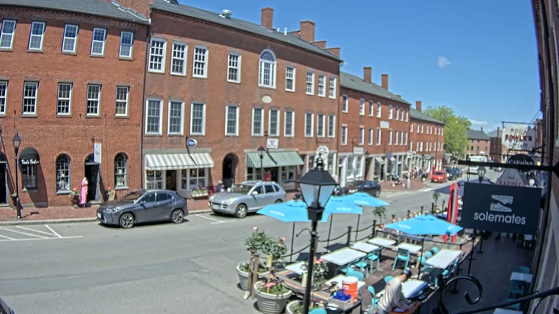 A sunny day on a brick-lined street with parked cars, outdoor seating at a restaurant, and several shops with awnings.