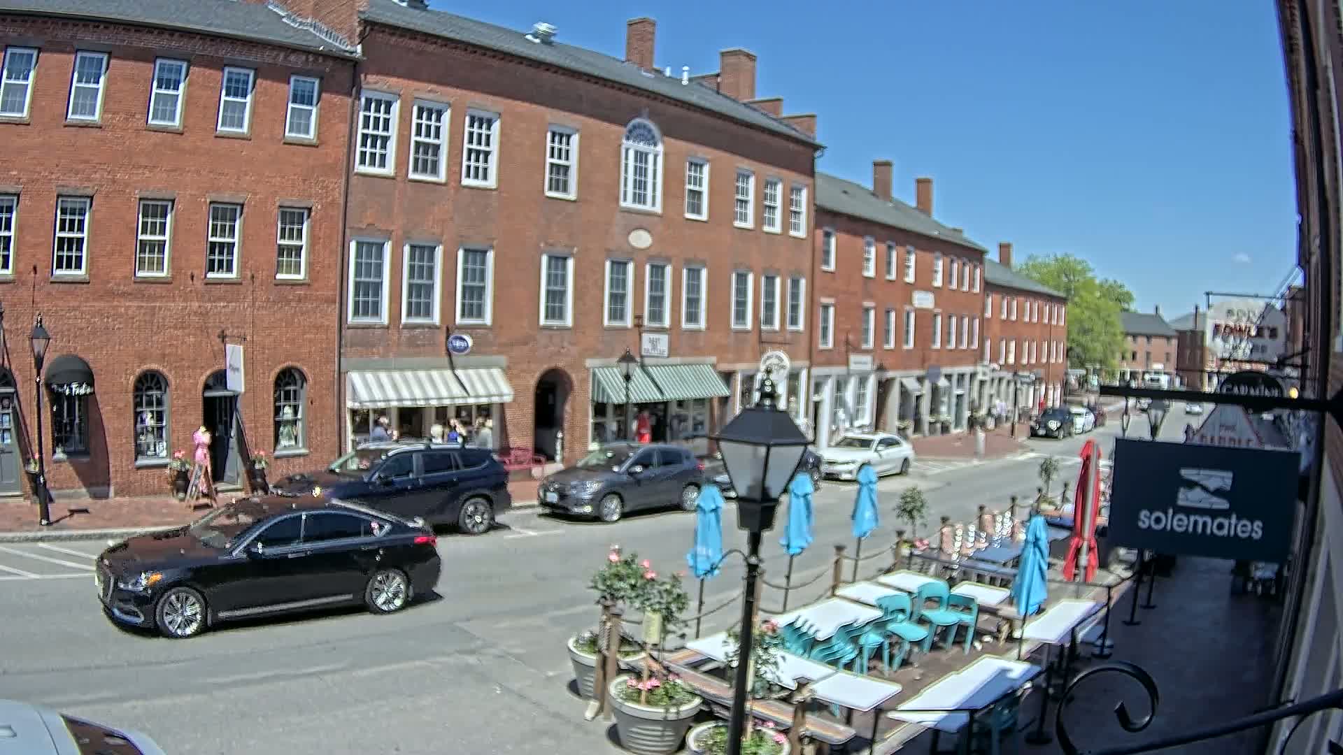 A sunny day on a brick-lined street with shops, parked cars, and an outdoor patio seating area with teal chairs and umbrellas.