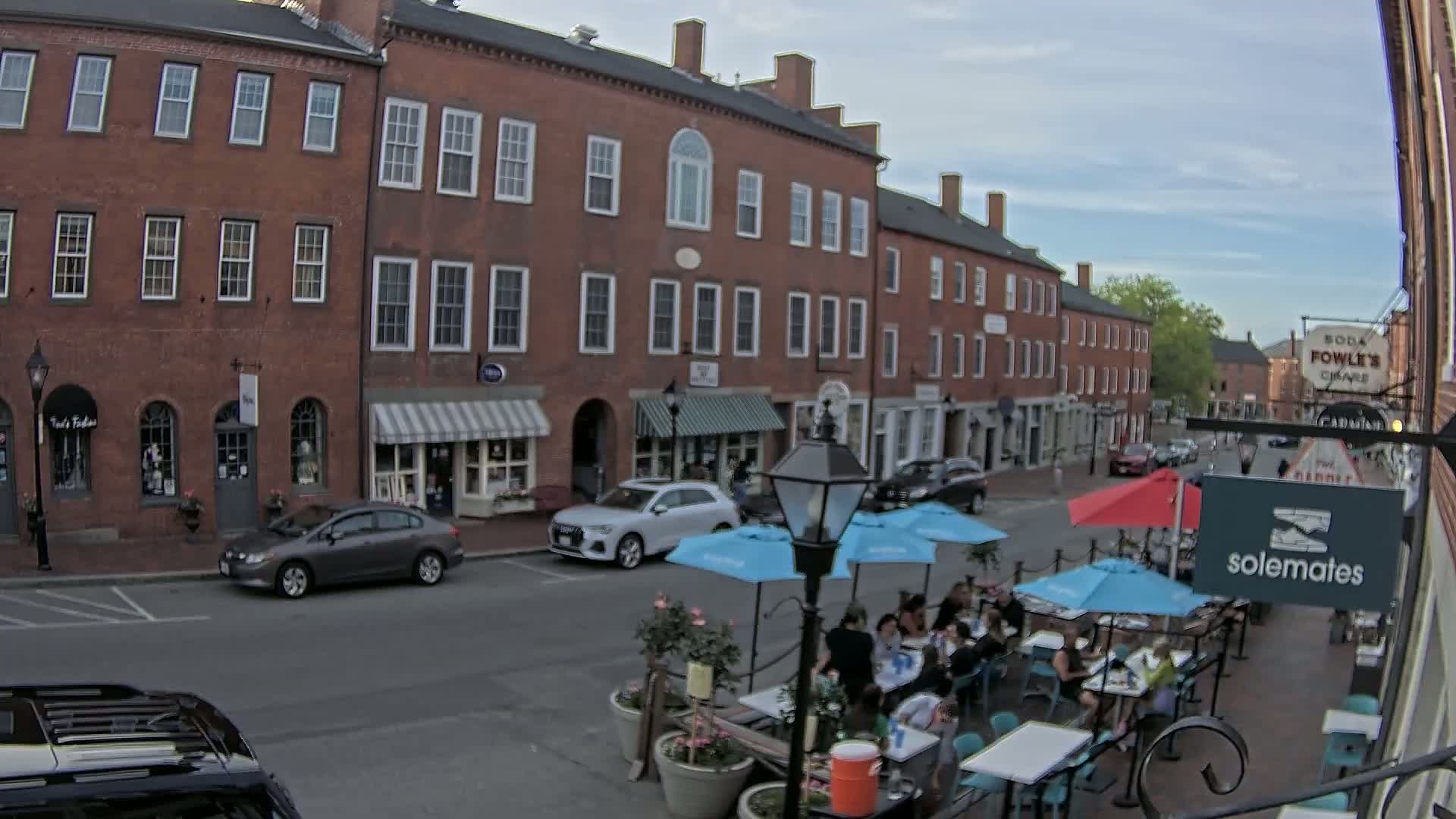 A brick street lined with shops and restaurants features outdoor seating under umbrellas on a partly cloudy day.