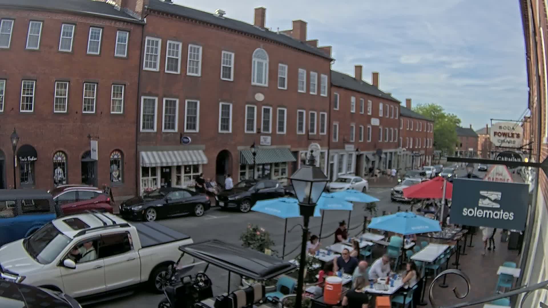A brick street with shops and restaurants is bustling with activity under a partly cloudy sky, with people dining outdoors at tables under umbrellas.