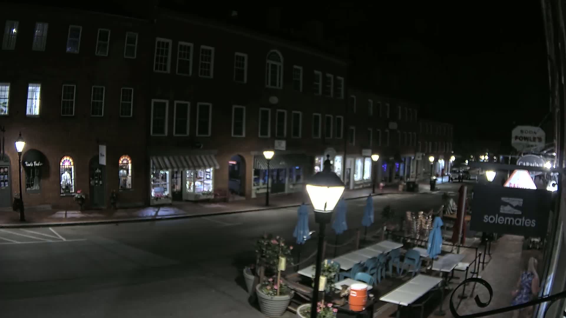 A nighttime street scene shows a row of brick buildings with shops on the ground floor, outdoor seating with teal chairs and tables, and lampposts illuminating the empty street.