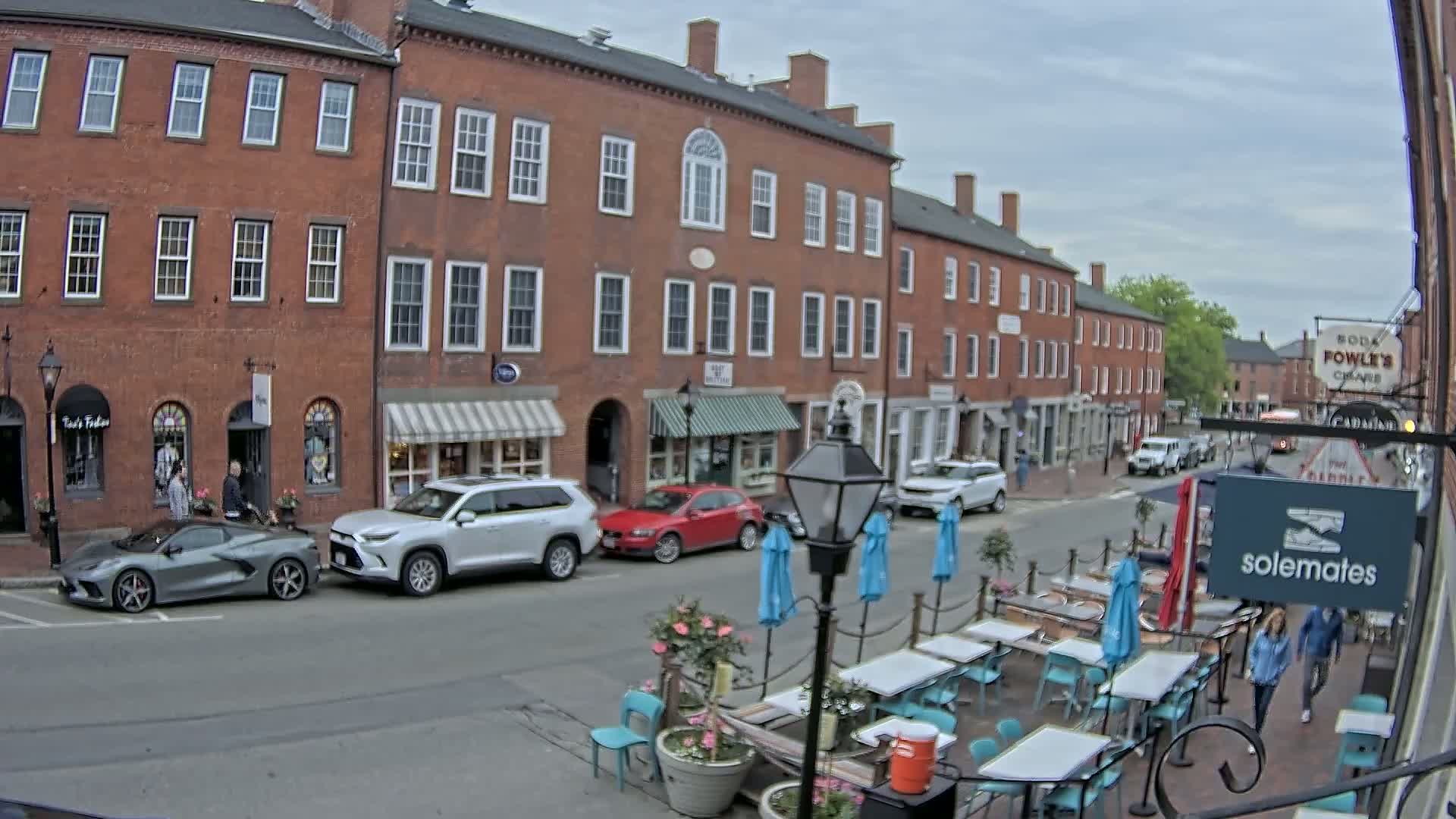 A street scene on an overcast day shows a row of brick buildings with shops and restaurants, cars parked along the street, and an outdoor patio seating area with tables and umbrellas.