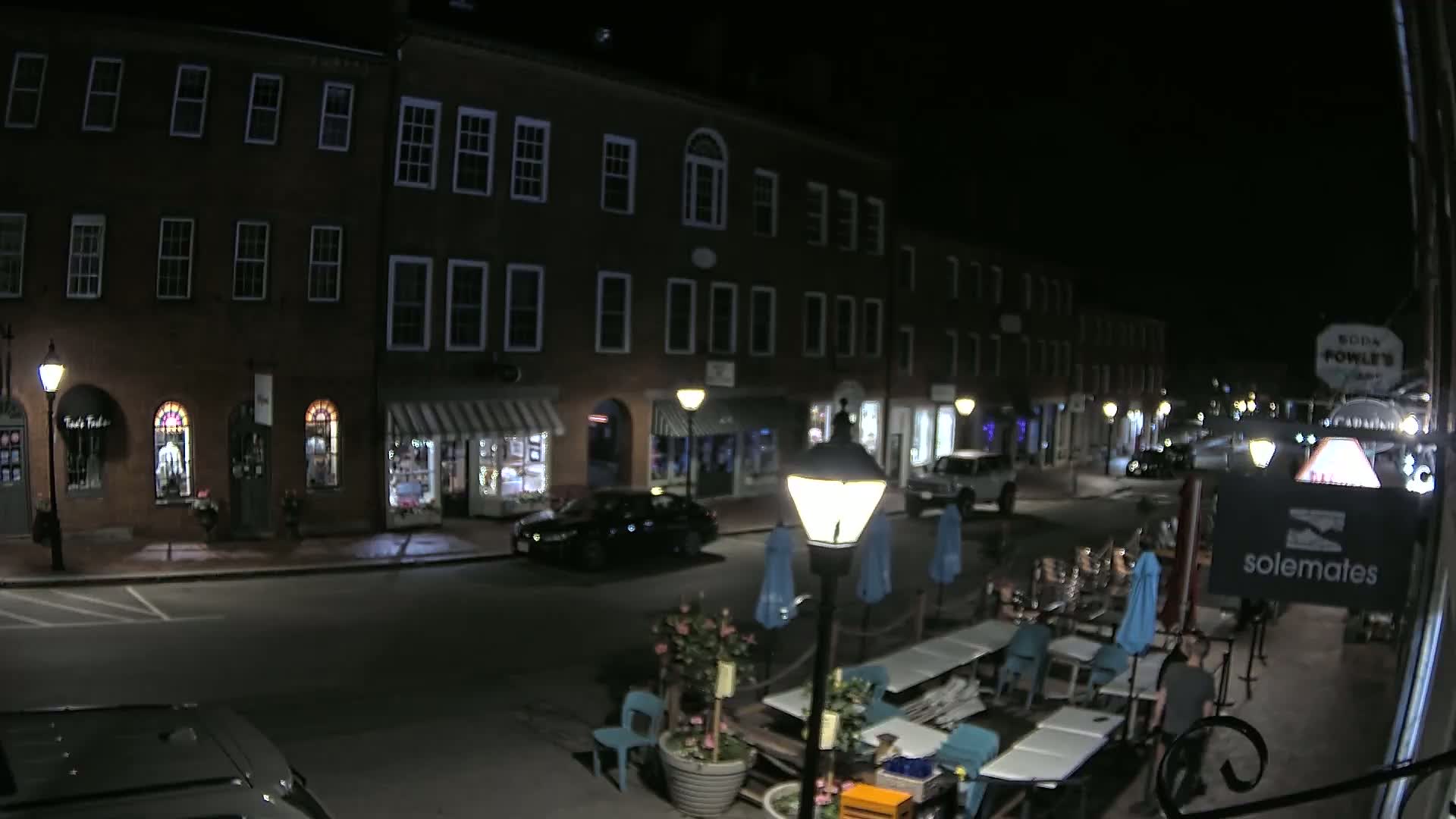 A nighttime street scene shows a row of brick buildings with shops and restaurants, outdoor seating, and a few cars parked along the side of the road.