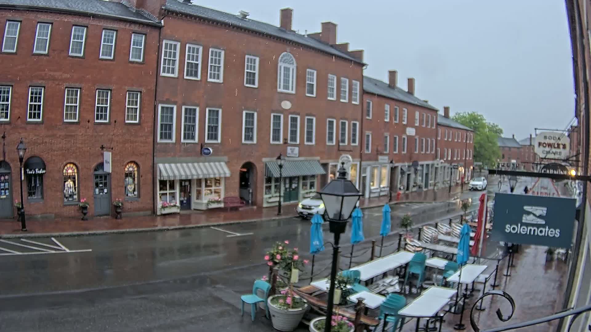 A rain-slicked street in front of brick buildings with outdoor seating and umbrellas under a gray sky.