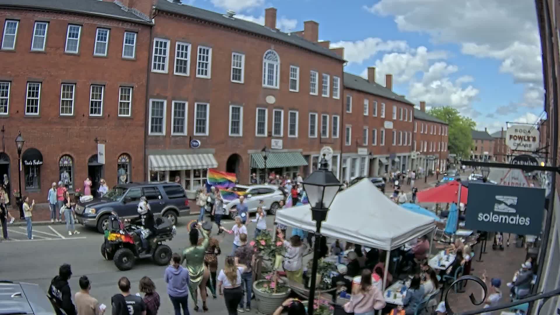 A street scene on a partly cloudy day shows a crowd of people gathered outdoors near brick buildings, some under a tent, with a person on an ATV and a rainbow flag visible.
