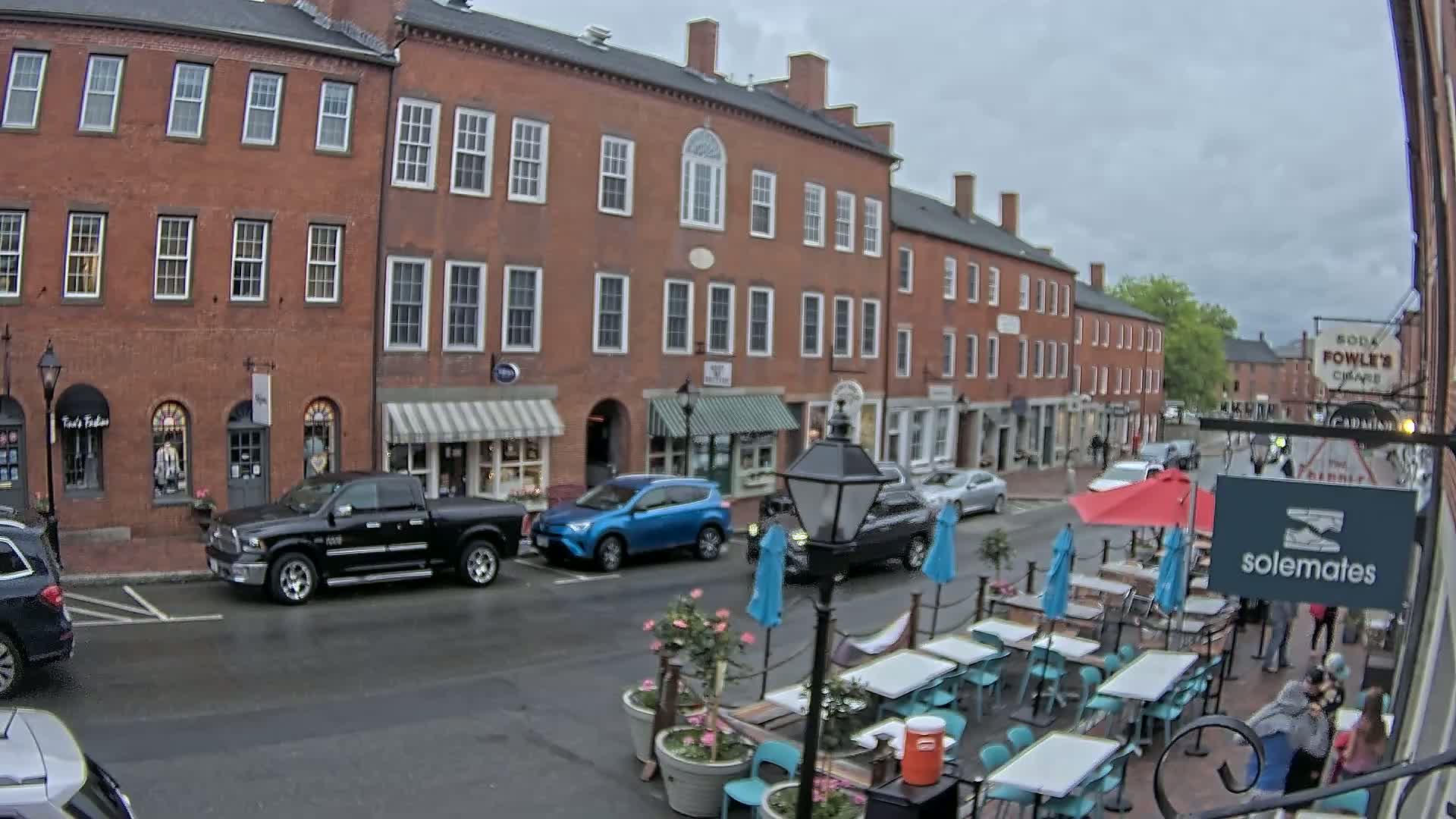 A rain-slicked street in front of brick buildings with shops and an outdoor cafe with teal chairs and tables under umbrellas.