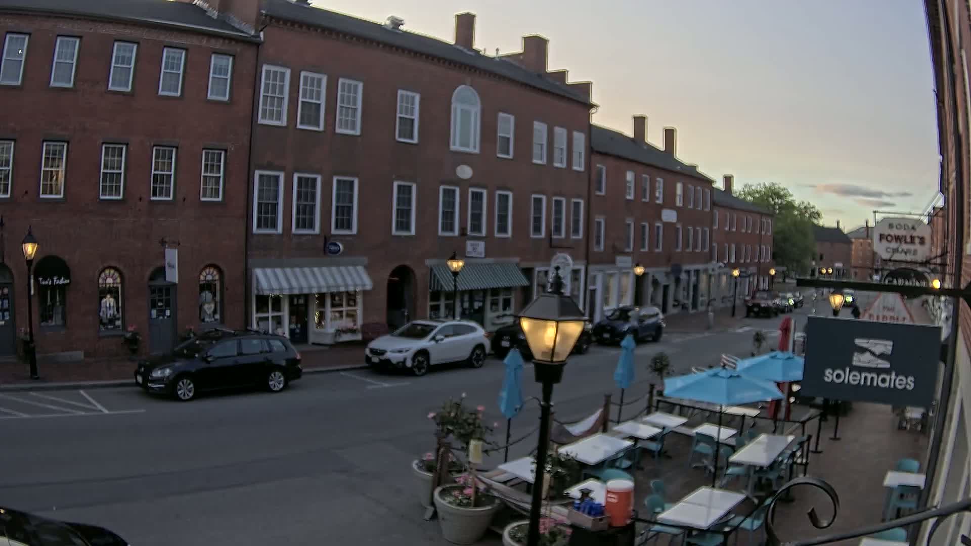 A street scene at dusk shows a row of brick buildings with shops and outdoor seating at a restaurant.