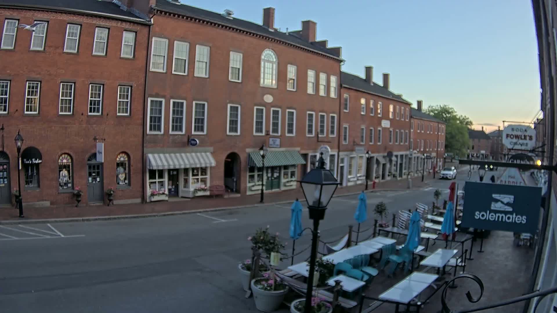 A mostly empty street lined with brick buildings and shops, featuring outdoor seating at a cafe under a clear evening sky.