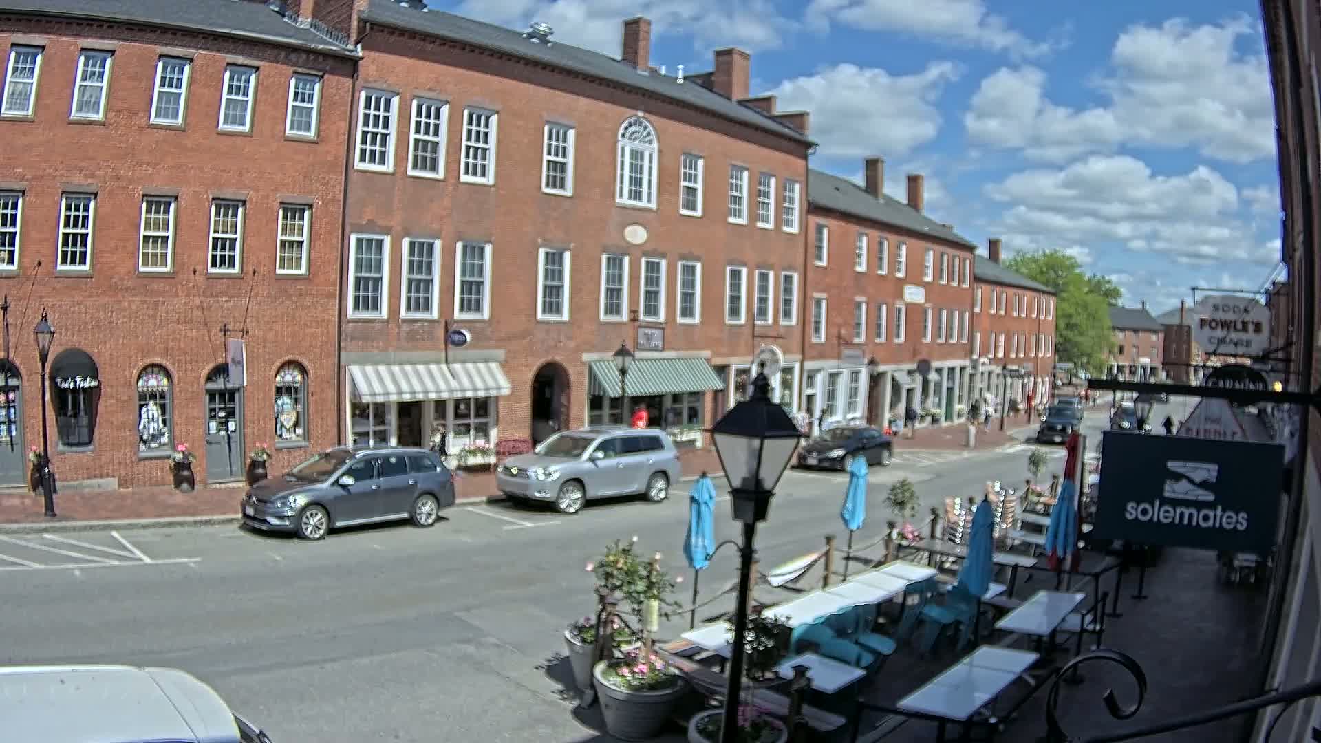 A sunny day on a brick-lined street shows several parked cars in front of shops with outdoor seating.