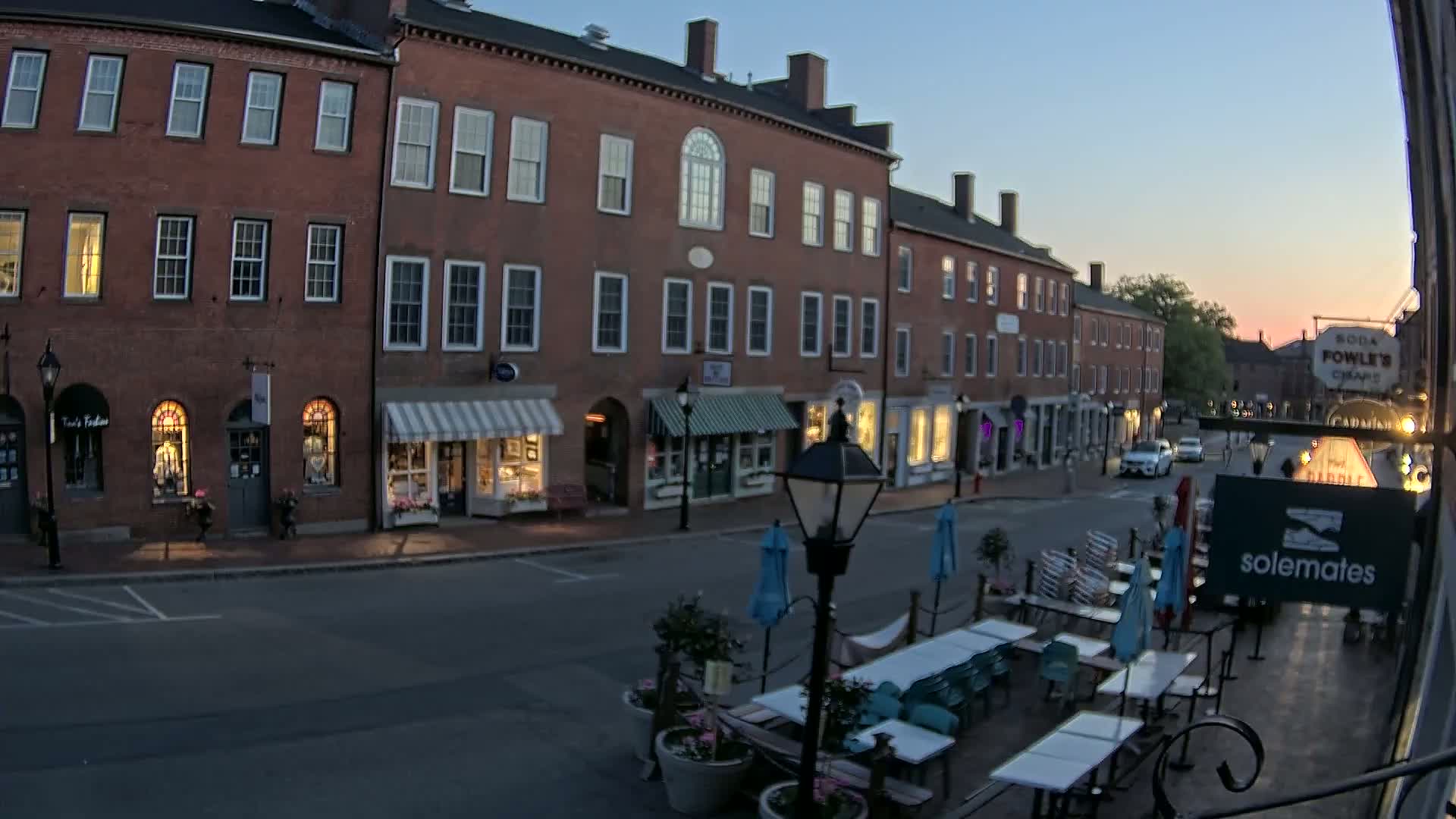 A twilight view of a brick-lined street with shops and outdoor seating, under a clear, dusky sky.
