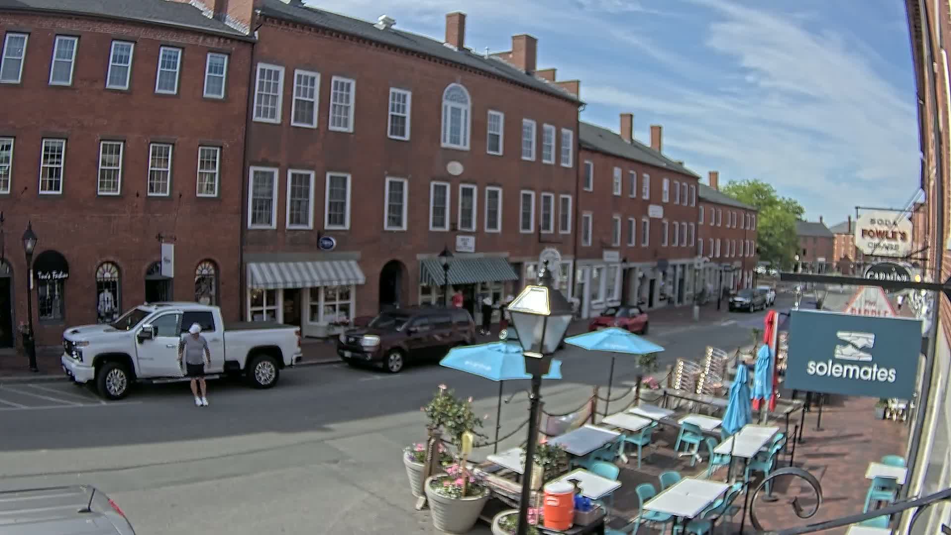 A street scene on a sunny day shows brick buildings with shops and a restaurant patio with turquoise tables and umbrellas, and a white pickup truck parked nearby.