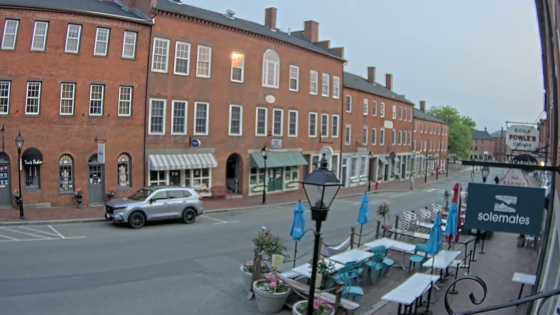 A silver SUV is parked on a street in front of several brick buildings with shops and outdoor seating under blue umbrellas on a cloudy day.