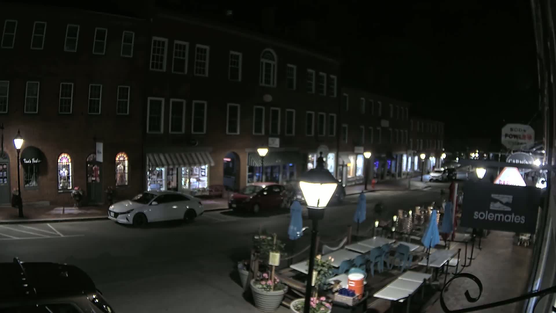 A nighttime street scene shows a row of brick buildings with shops and a few parked cars, and an outdoor seating area with tables and umbrellas in the foreground.