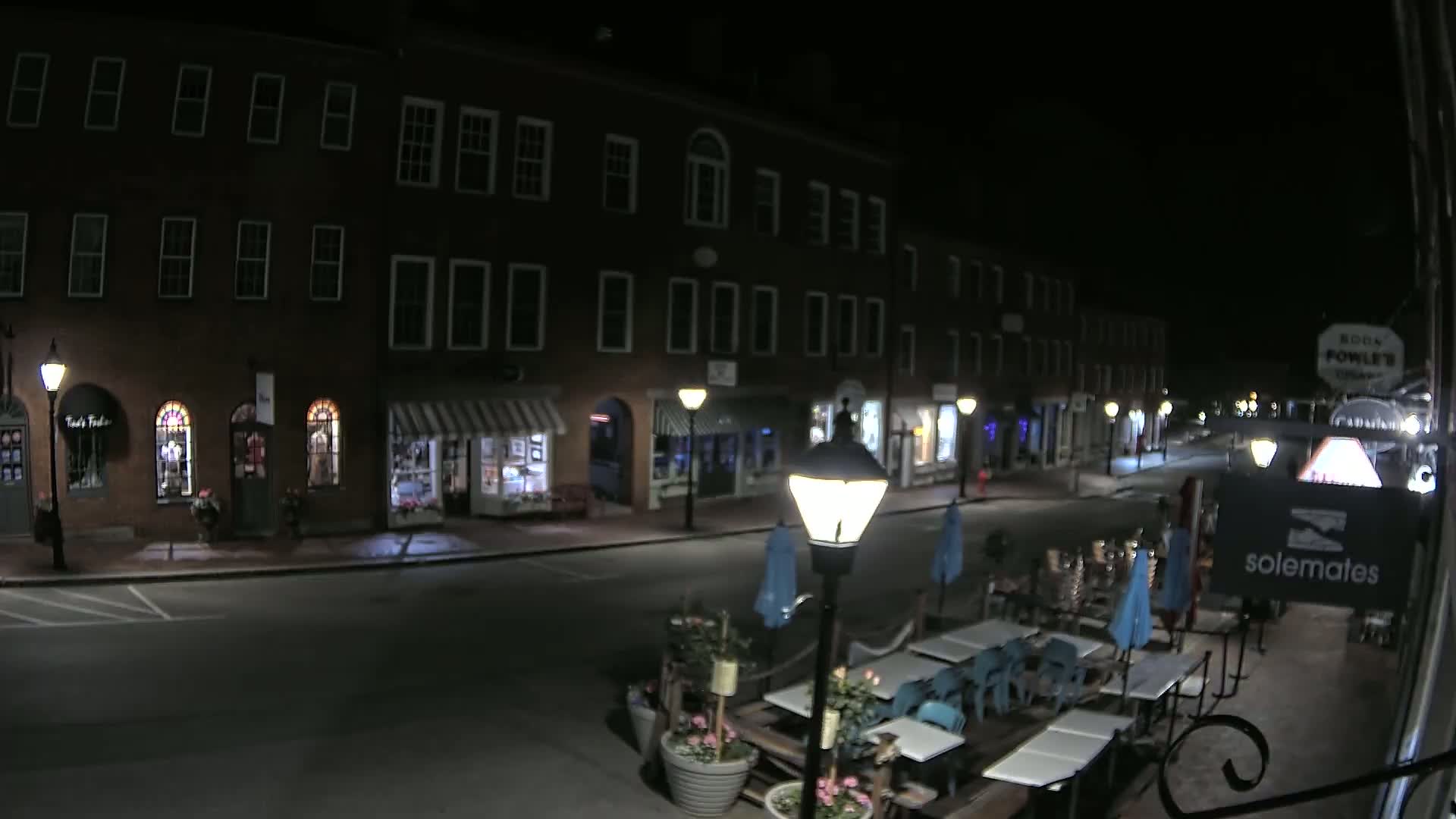 A nighttime street scene shows a row of brick buildings with storefronts, outdoor cafe seating, and streetlights illuminating a mostly empty road.