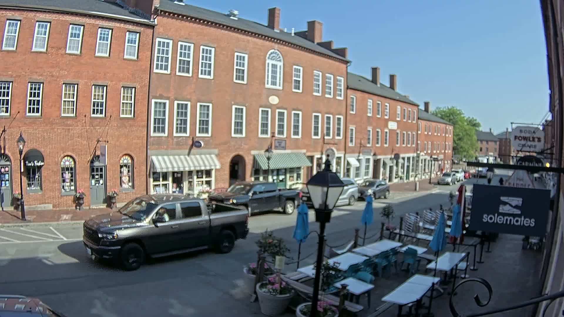 A sunny day on a brick-lined street with parked cars, a pickup truck driving by, and outdoor seating at a restaurant.