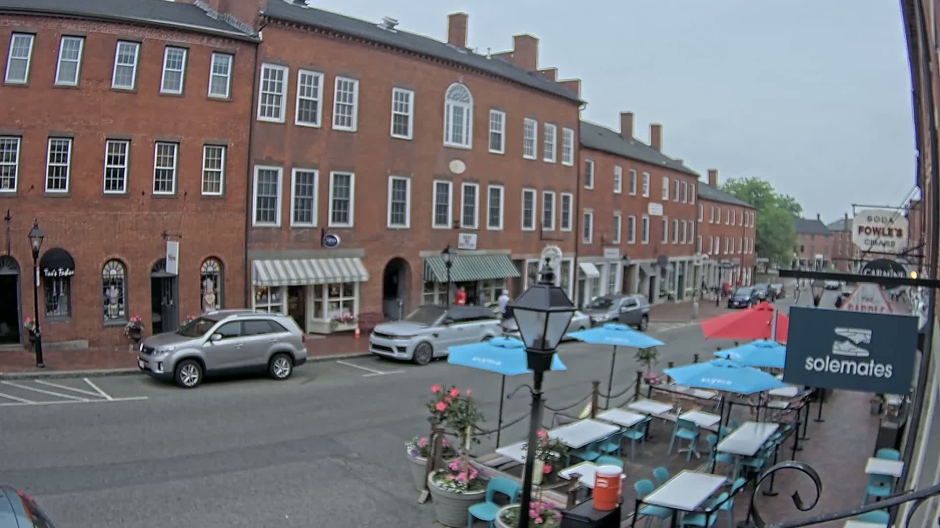 A street scene on an overcast day shows several brick buildings with shops and a patio dining area with light blue umbrellas and turquoise chairs.