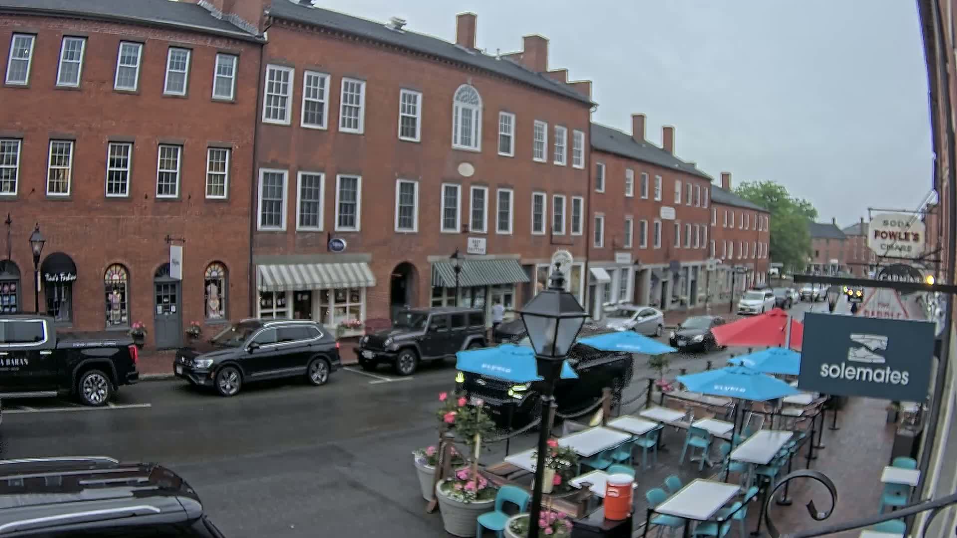A brick street, lined with shops and restaurants, has outdoor seating under blue and red umbrellas on a cloudy day.