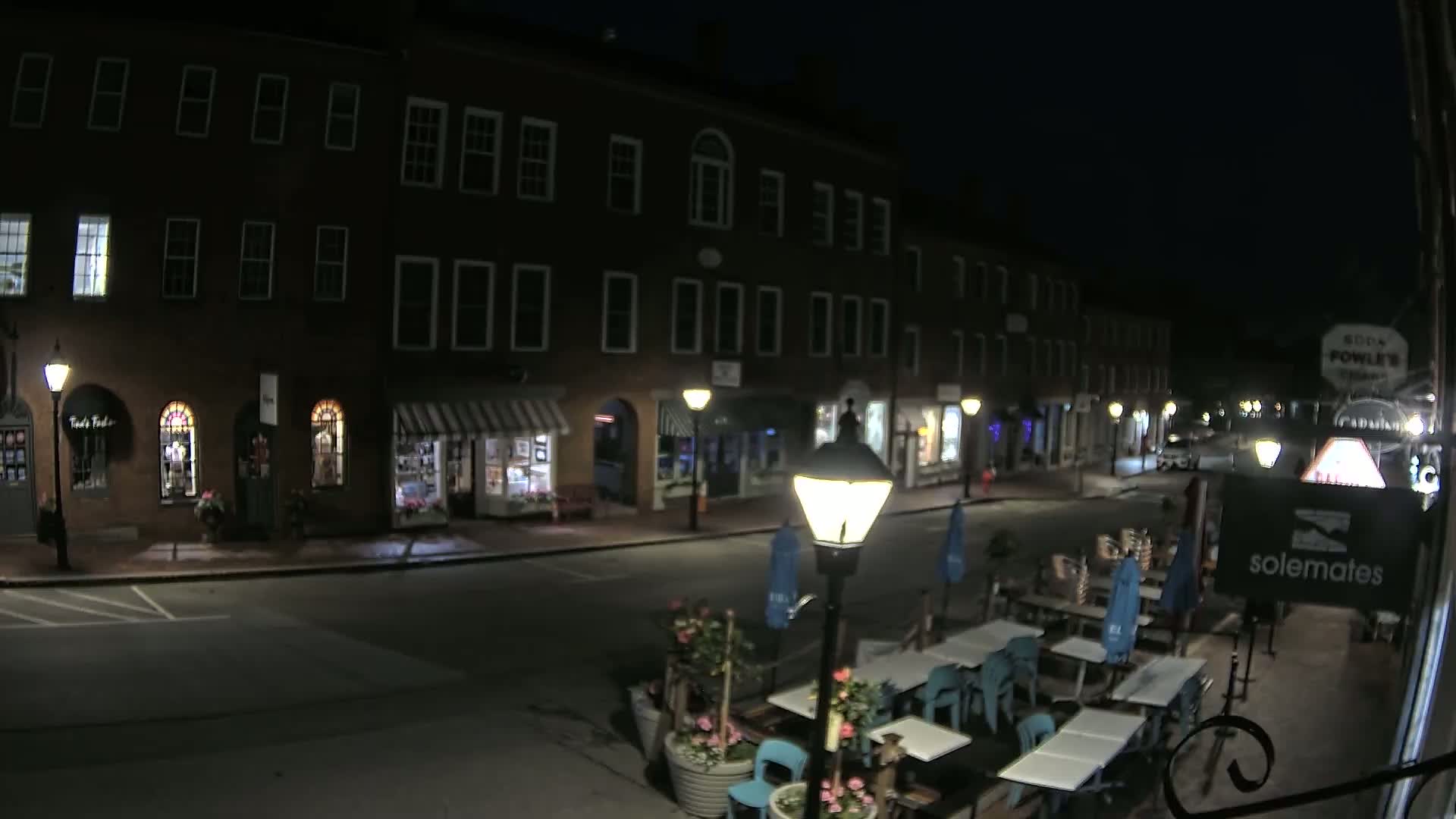 A nighttime street scene shows a row of brick buildings with shops and outdoor seating, under a dark sky.