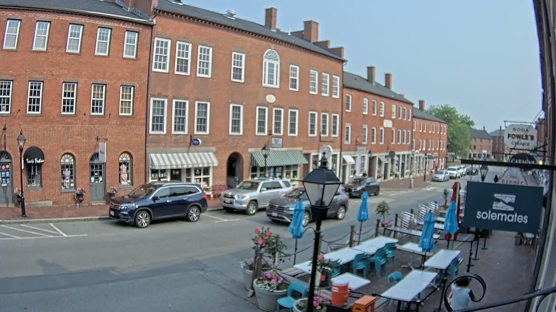 A street scene on a sunny day shows several brick buildings with storefronts, parked cars, and an outdoor cafe with blue umbrellas and turquoise chairs.