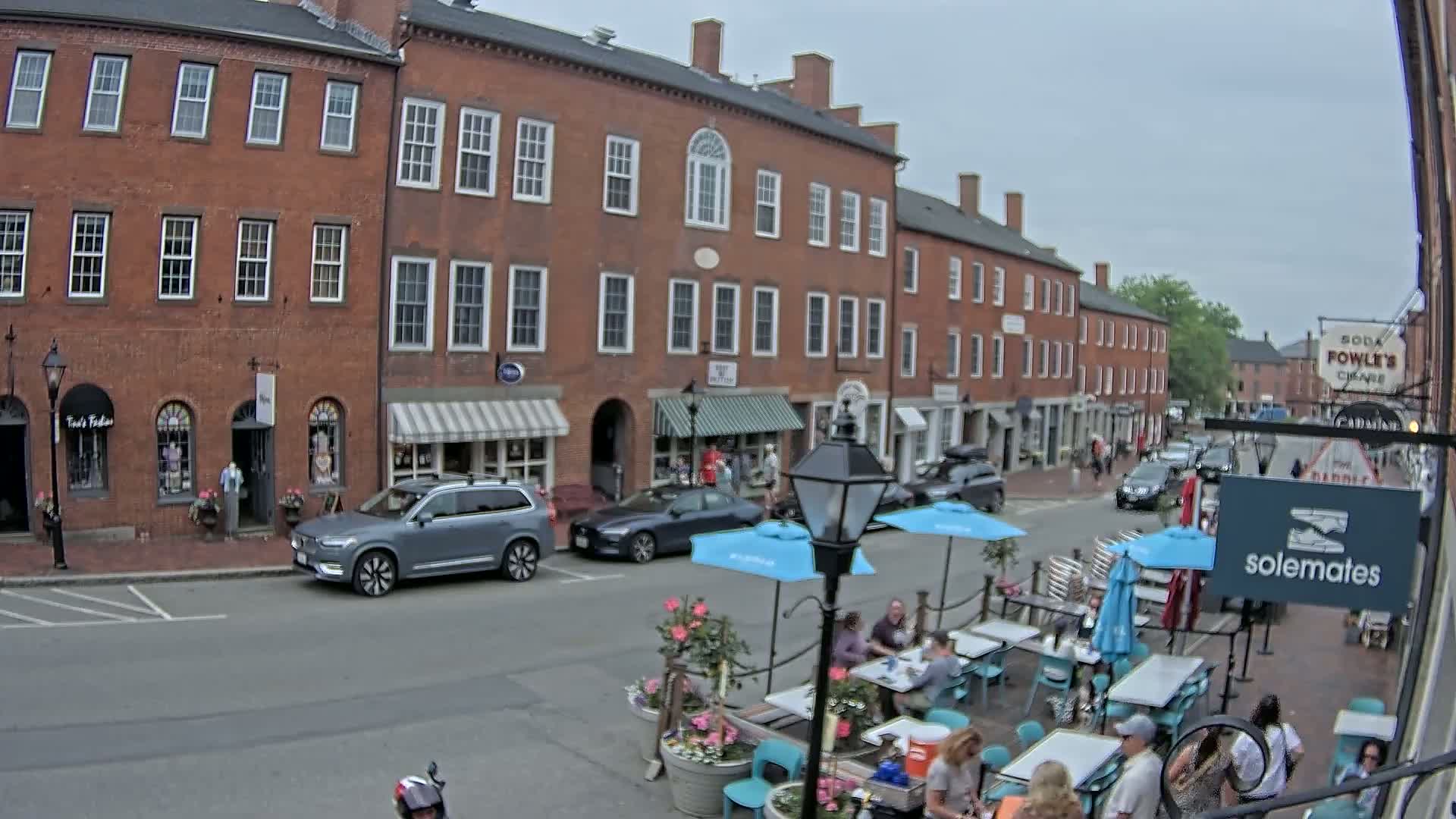 A street scene on an overcast day shows several brick buildings with shops and outdoor seating at a cafe, with cars parked along the street.