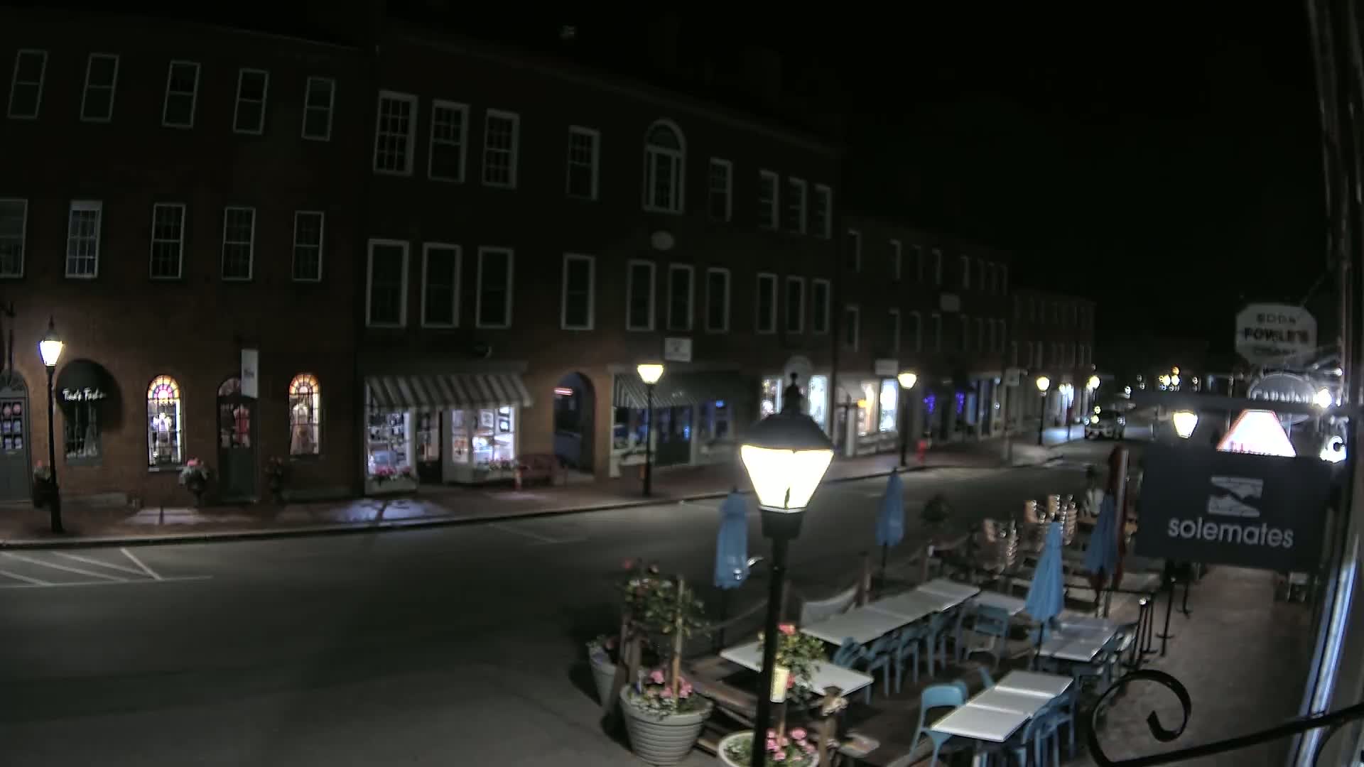 A nighttime street scene shows several brick buildings with shops and a restaurant's outdoor seating area with tables and chairs.