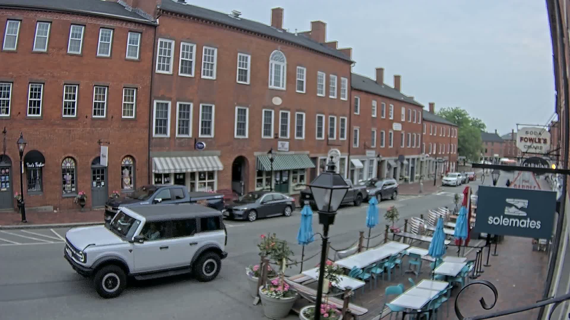 A street scene shows a row of brick buildings with shops and outdoor seating under blue umbrellas, a light-grey SUV driving down the street, and overcast skies.