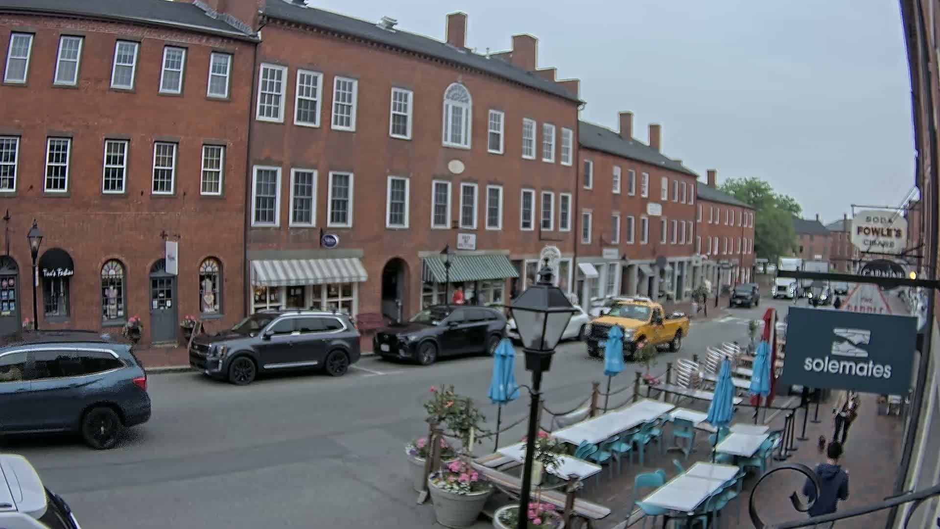 A street scene on an overcast day shows several parked cars in front of brick buildings with shops and awnings, and an outdoor seating area with tables and umbrellas.