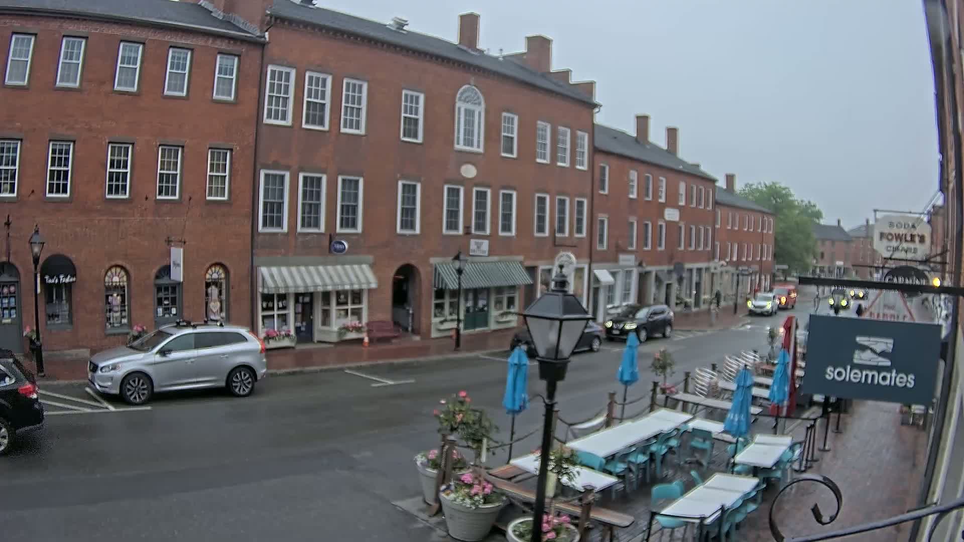 A rain-slicked street in front of a row of brick buildings with outdoor seating and parked cars.