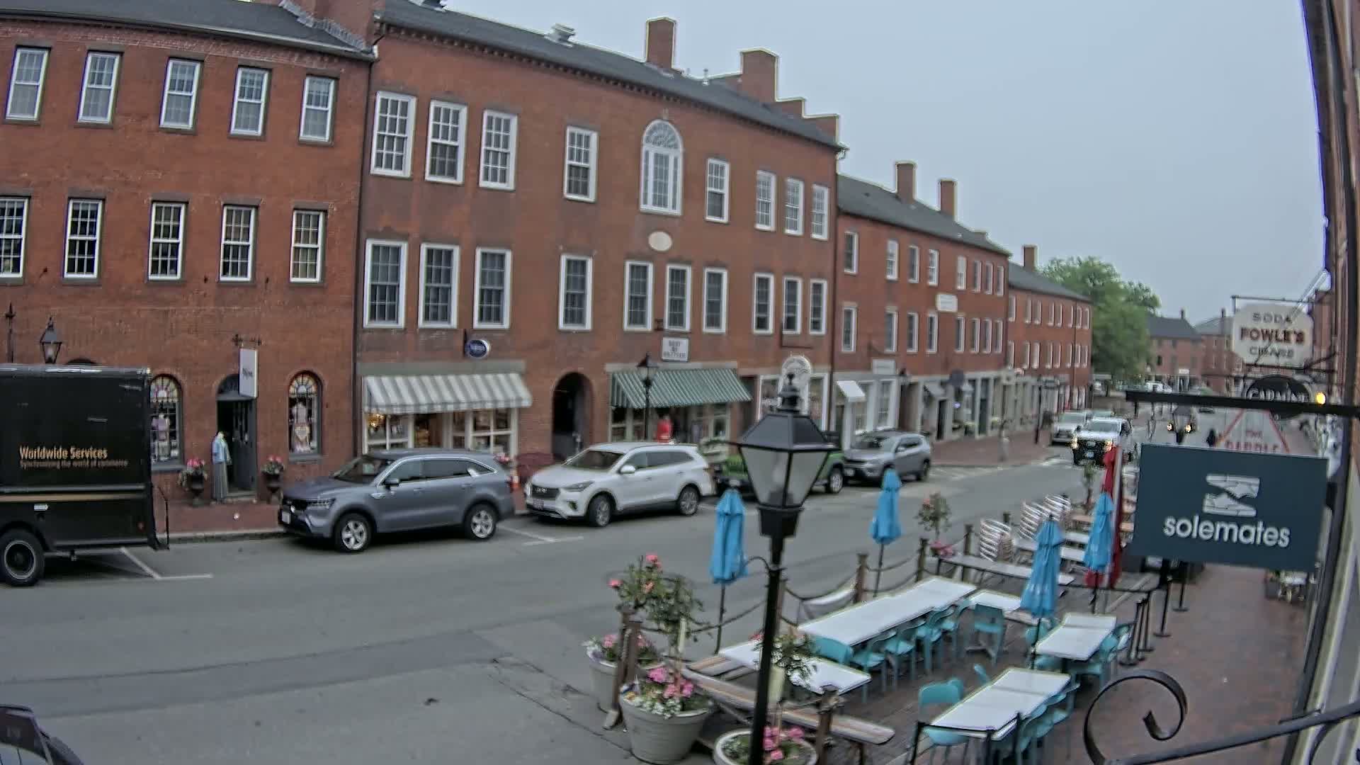 A brick street with parked cars and an outdoor cafe under an overcast sky.