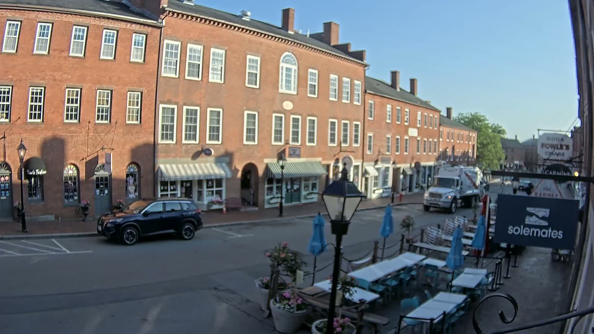 A sunny day on a brick-lined street shows a row of shops and restaurants with outdoor seating, a parked SUV, and a delivery truck driving away.