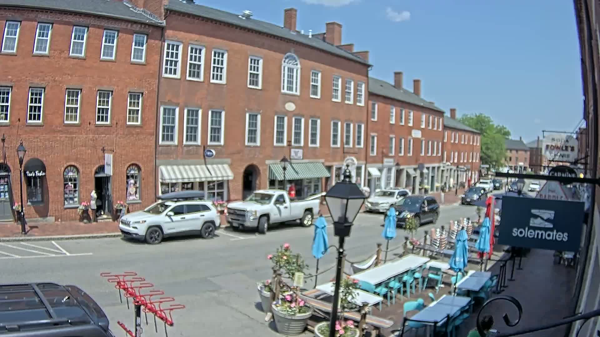A sunny day reveals a brick street lined with shops and a few parked cars, with outdoor seating at a restaurant in the foreground.