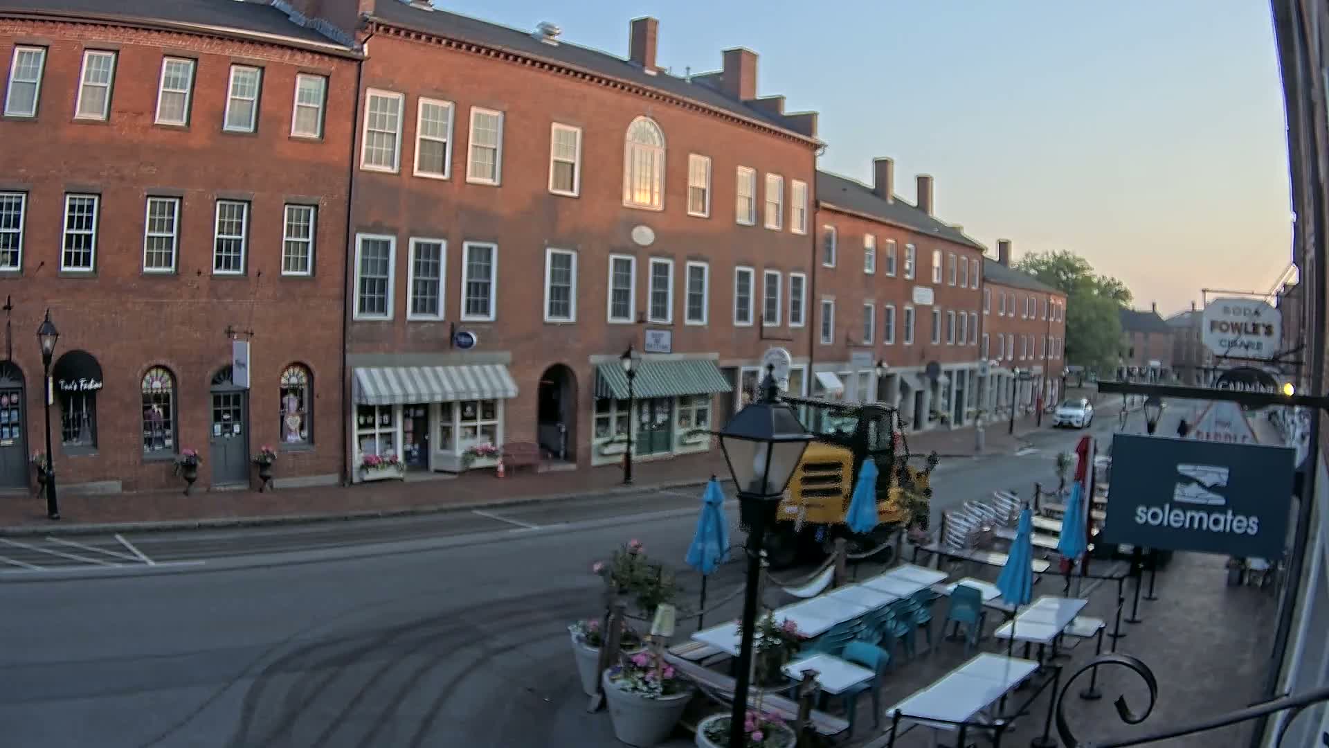 A brick street lined with shops and a streetside patio with teal chairs and umbrellas under a clear, early morning sky.