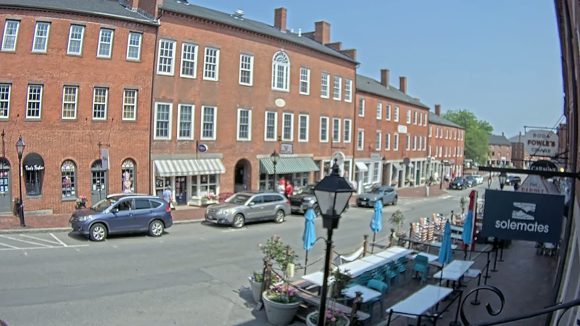 A sunny day on a brick-lined street shows several shops and restaurants with cars parked along the side, and outdoor seating with turquoise chairs and umbrellas in front of one establishment.