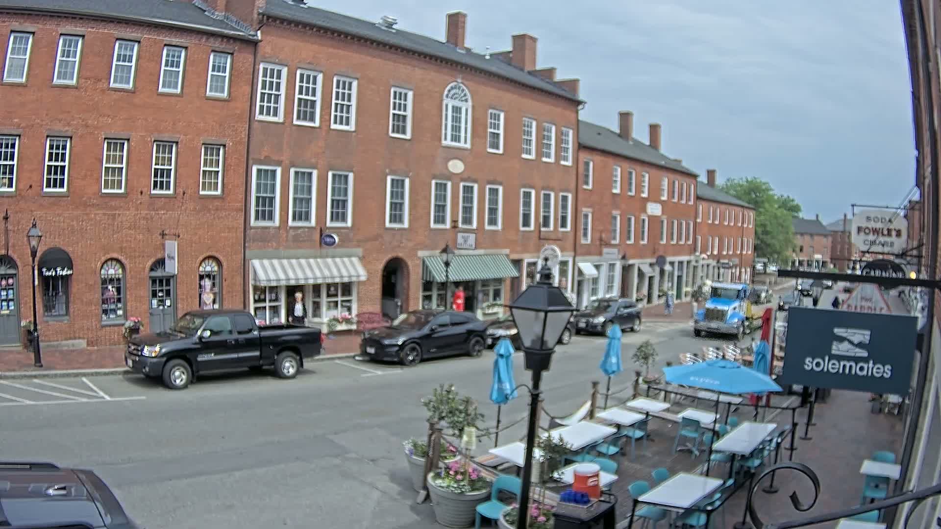 A street scene on an overcast day shows several parked cars in front of brick buildings with shops and restaurants, one of which has outdoor seating with teal tables and chairs under blue umbrellas.