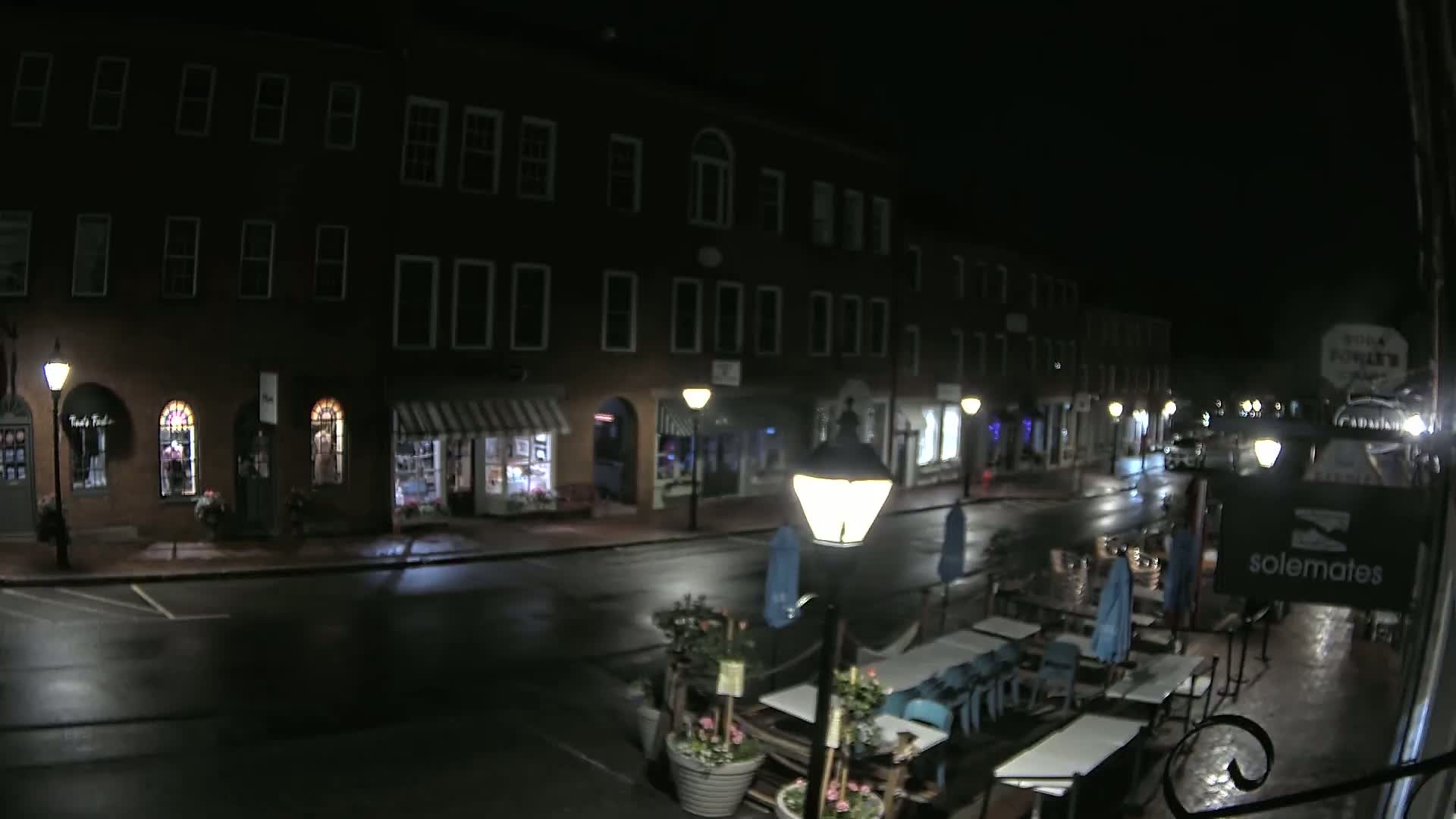 A wet, dark street at night lined with shops and buildings, showing outdoor seating at a restaurant.