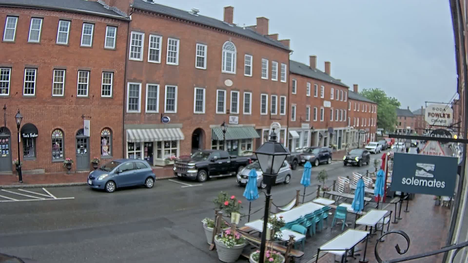 A rain-slicked street in front of several brick buildings with awnings and outdoor seating for a restaurant.
