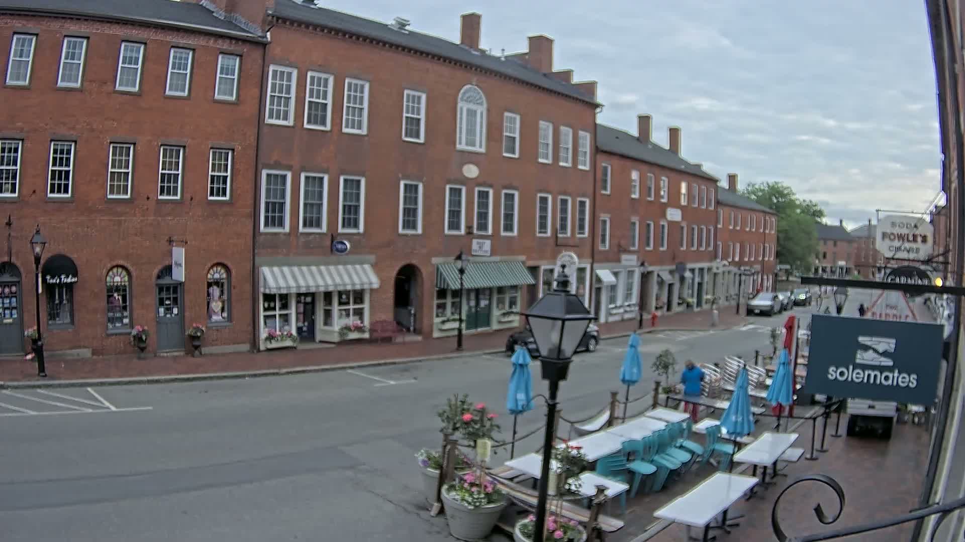 A street scene on an overcast day shows a row of brick buildings with shops on the ground floor, outdoor seating with teal chairs and tables, and a few cars parked on the street.