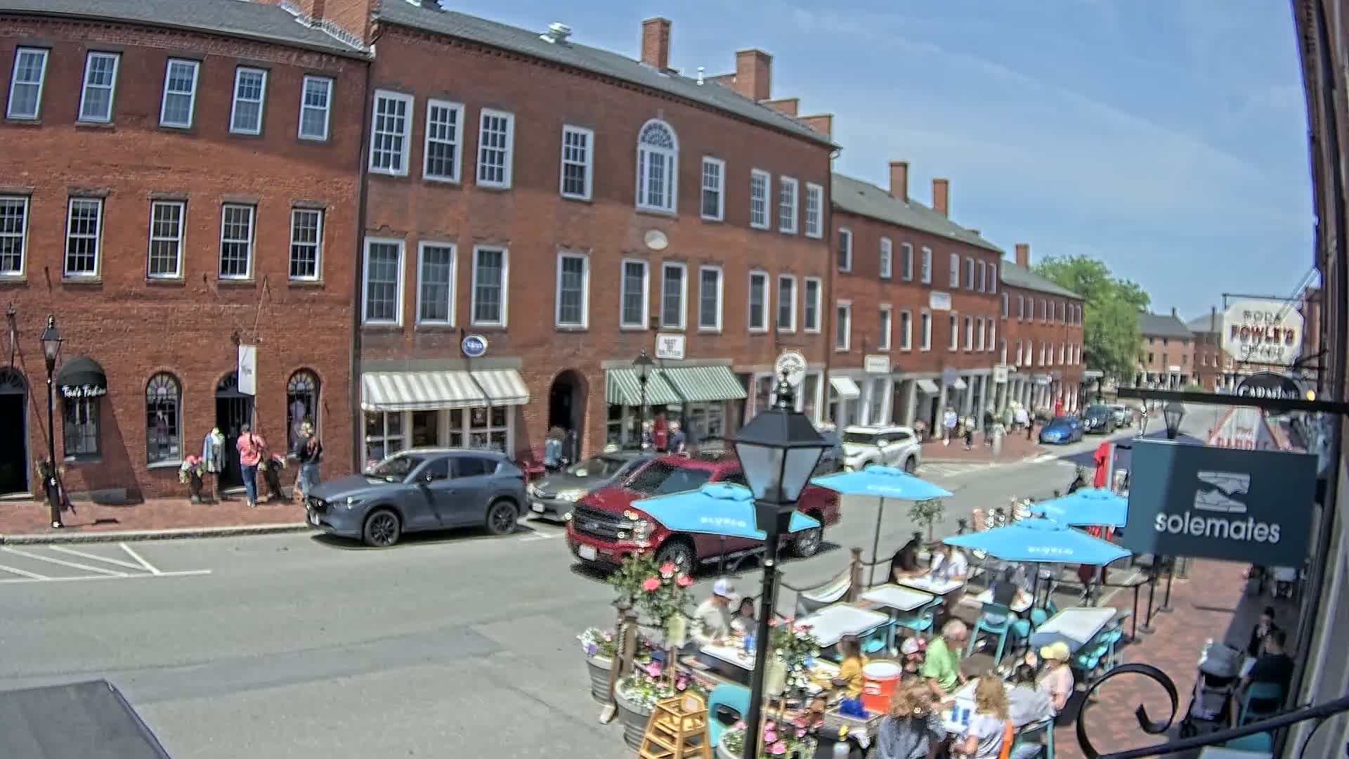 A sunny street scene shows a row of brick buildings with shops and outdoor seating, where people are eating at tables under blue umbrellas.