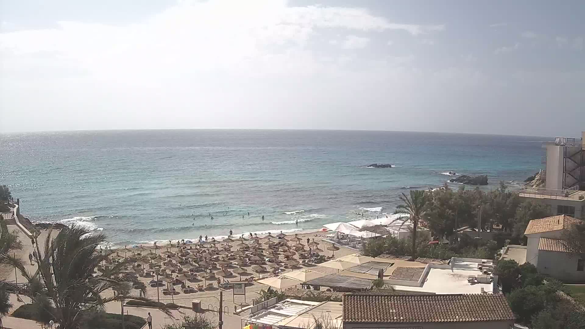 A mostly sunny day shows a sandy beach with many umbrellas and people, adjacent to calm, light-blue ocean water.