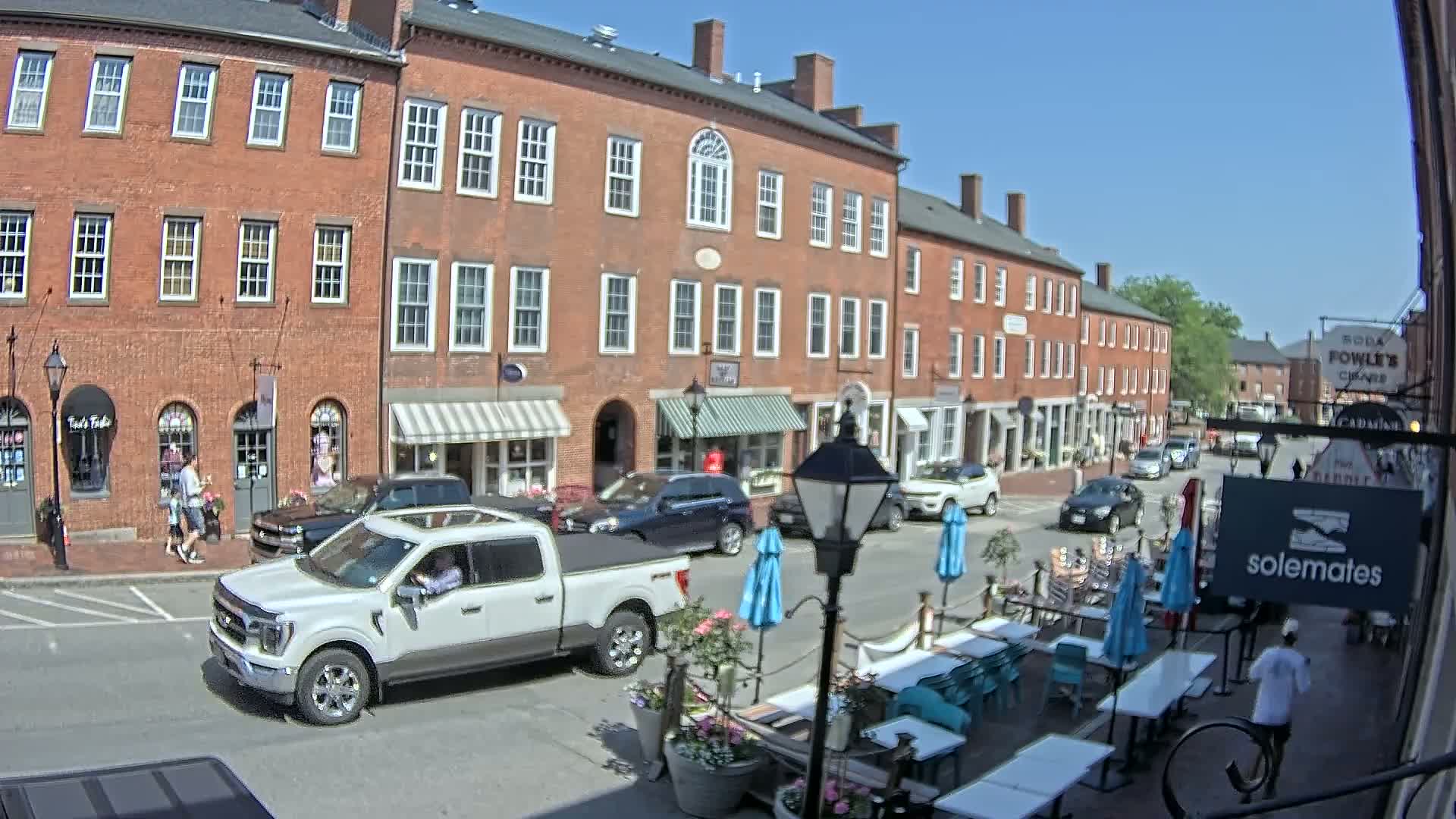 A sunny day on a brick-lined street shows a row of shops and restaurants with outdoor seating, including a white pickup truck parked in front of one of them.