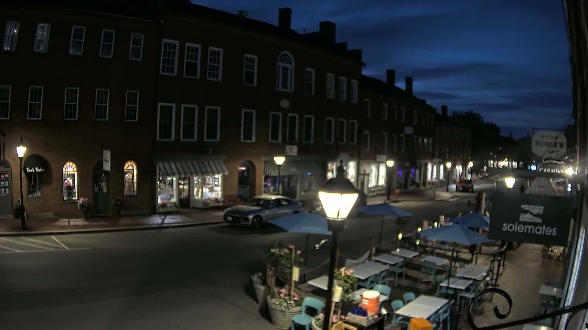 A nighttime street scene shows a row of brick buildings with outdoor seating at a restaurant under blue umbrellas, with a car parked on the street under a dark, mostly clear sky.