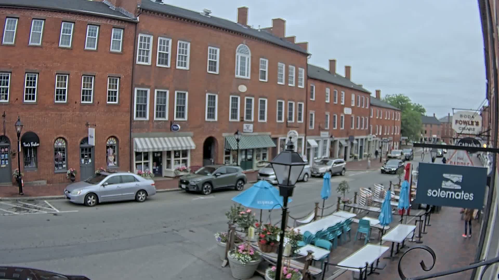 A street scene on an overcast day shows brick buildings with shops and outdoor seating, cars parked along the street, and pedestrians walking.