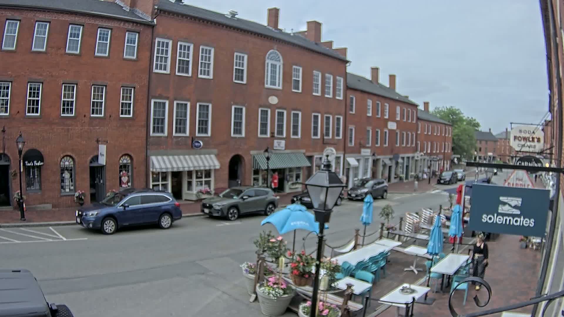 A street scene on an overcast day shows several brick buildings with shops on the ground floor and outdoor seating at a cafe in front of one.