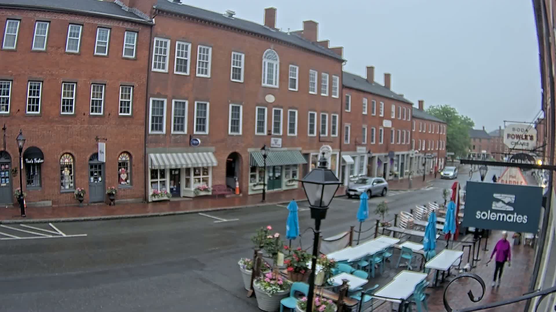 A wet brick street lined with shops and restaurants, featuring outdoor seating under umbrellas, on an overcast day.