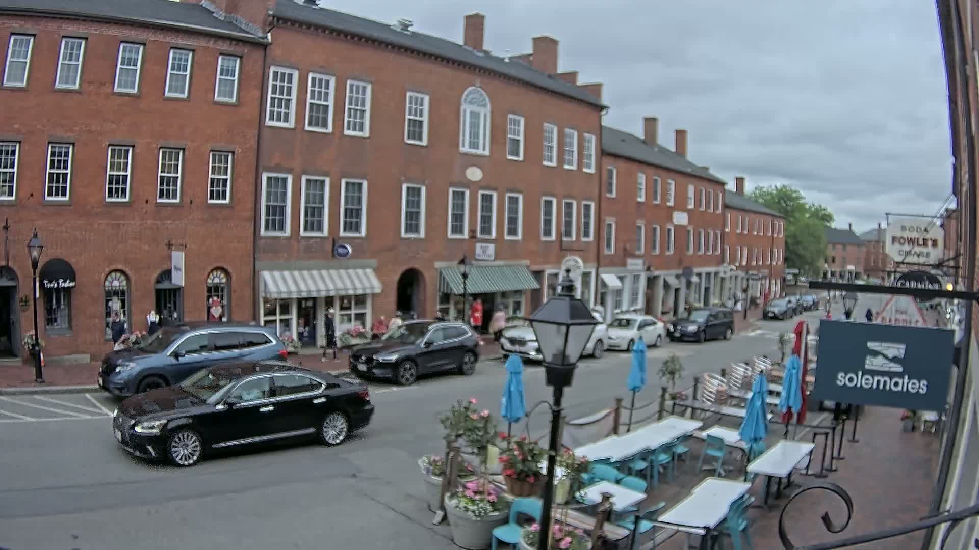 A street scene on an overcast day shows several brick buildings with shops on the ground floor, cars parked along the street, and outdoor seating with teal chairs and umbrellas in front of a restaurant.
