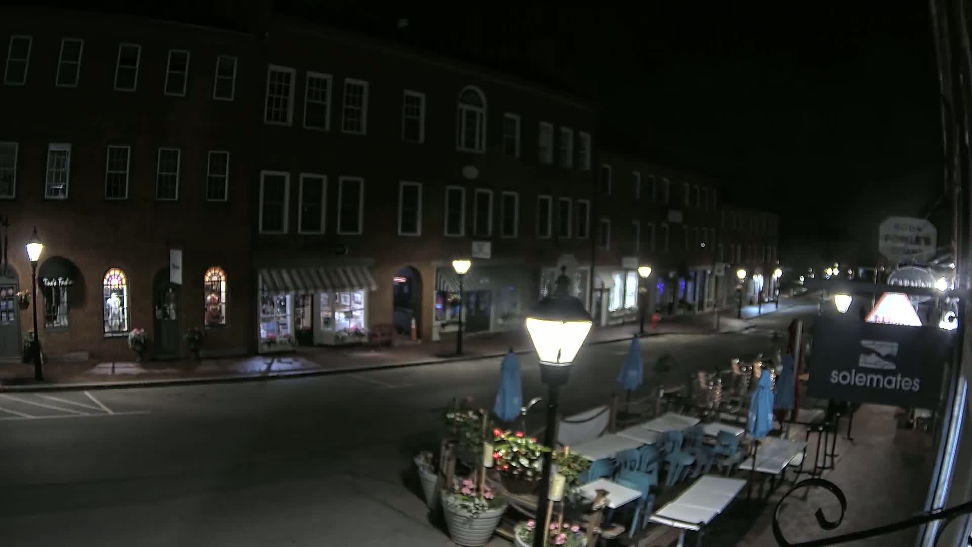 A nighttime street scene shows a row of brick buildings with shops and outdoor seating at a restaurant under lamplight.