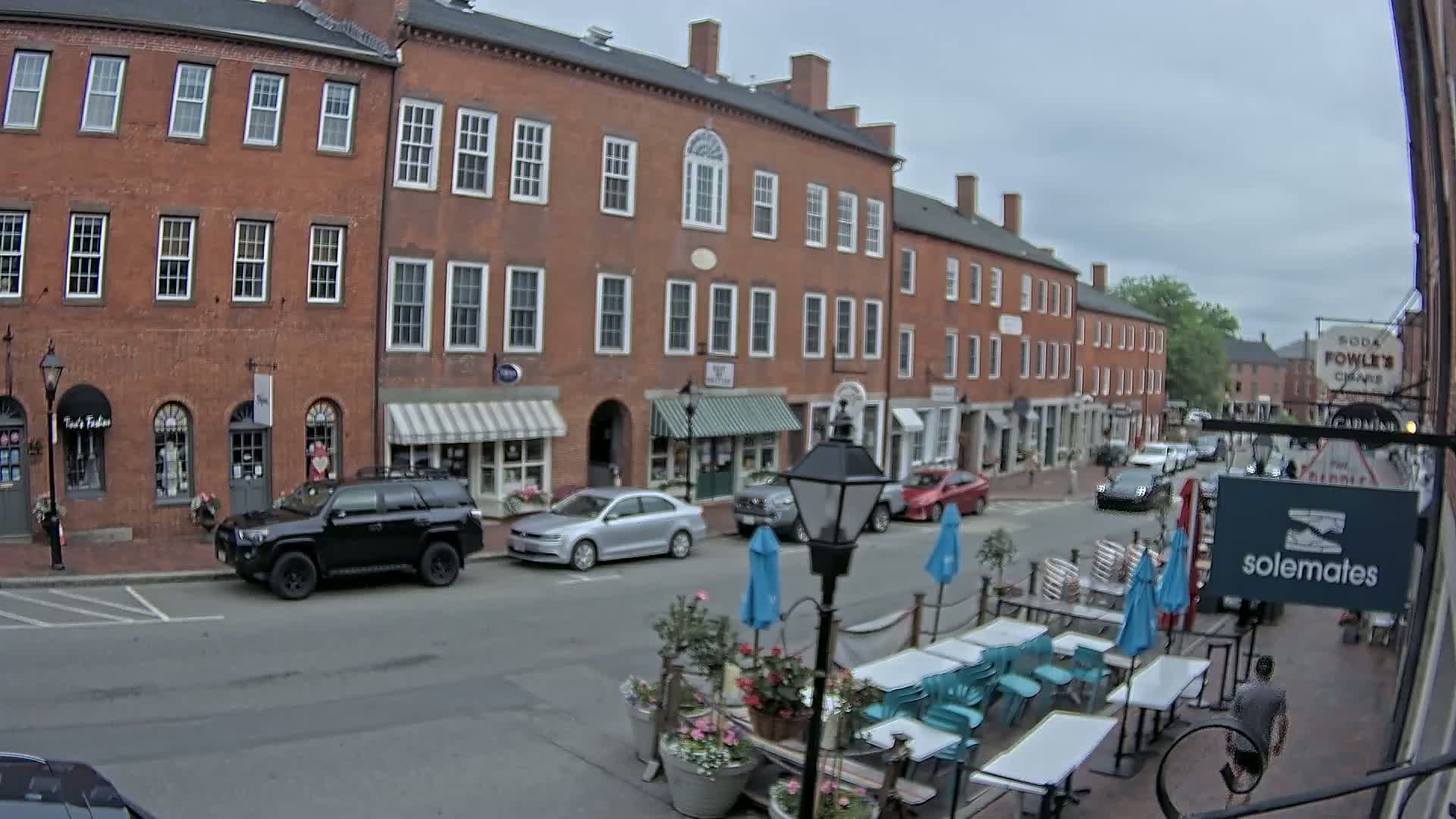 A street scene on an overcast day shows several brick buildings with shops on the ground floor, cars parked along the street, and outdoor seating at a cafe with blue umbrellas.