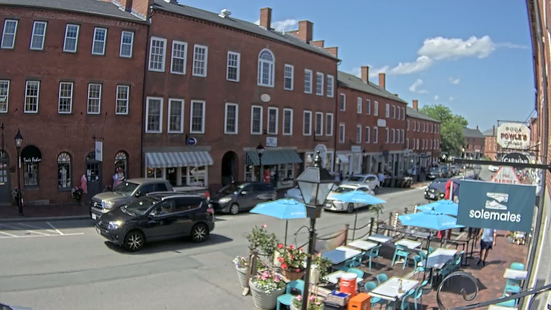 A sunny day on a brick-lined street with parked cars, outdoor seating, and several shops.
