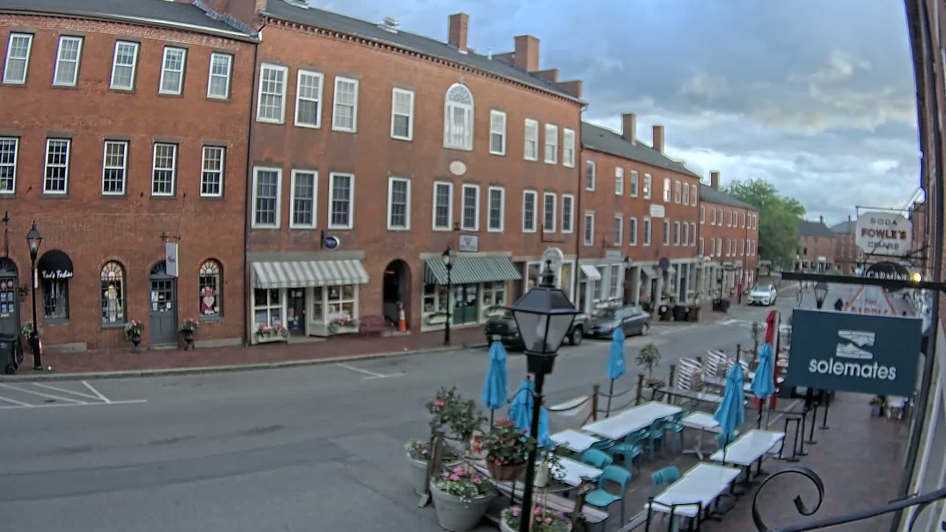 A street scene shows a row of brick buildings with shops and awnings, outdoor seating for a restaurant, and several cars parked along the street under a partly cloudy sky.